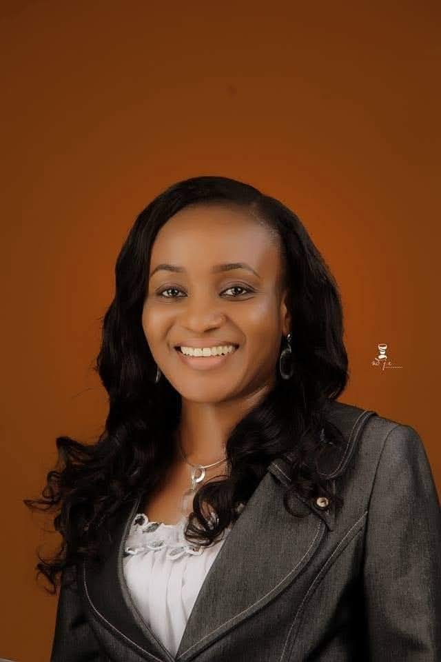A smiling woman with long, wavy hair, wearing a gray blazer and a white blouse, poses confidently against a warm brown background.
